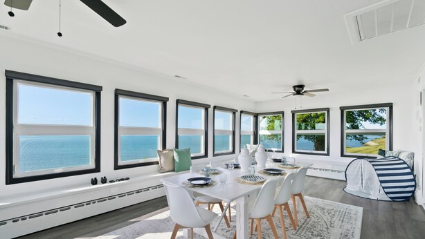 A dining area with a white table set for six, large windows with ocean views, a ceiling fan, and a striped play tent.