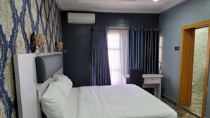 A hotel room with a bed, a desk, and a chair. There is a window with curtains, an air conditioning unit, and a door. - Camas Hotel and Suites (Abeokuta)