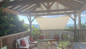 A covered outdoor seating area with a table and chairs, a sofa with cushions, and a view of the landscape.