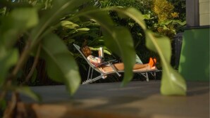 A person relaxing on a sun lounger in a garden area with lush greenery and a building in the background.