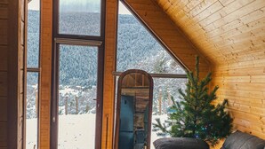En mysig stuga med träpanel i taket, ett stort fönster med utsikt över ett snötäckt berg, en liten julgran och en soffa i mörk färg.