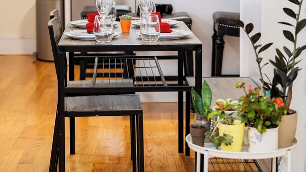 Un comedor con una mesa preparada para cuatro, una cocina al fondo y una planta en maceta sobre una mesita auxiliar.