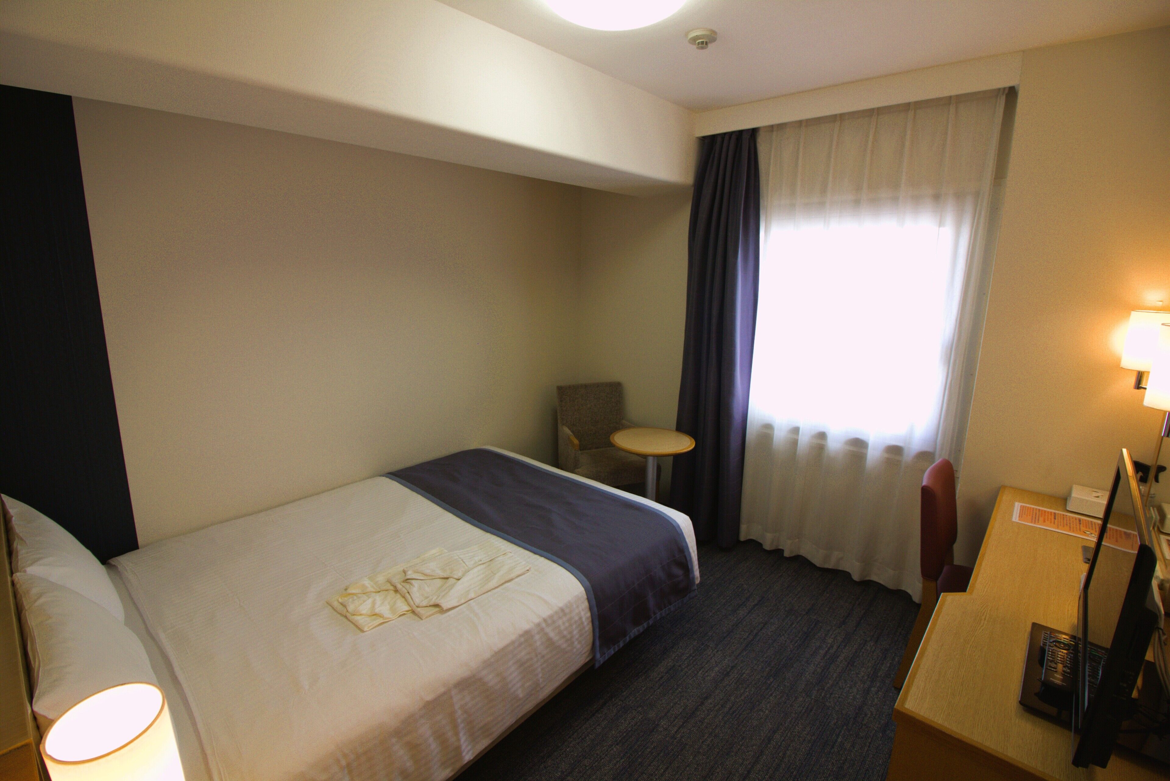Ein Hotelzimmer mit einem Einzelbett, einem kleinen Tisch, einem Stuhl und einem Schreibtisch mit Lampe.
