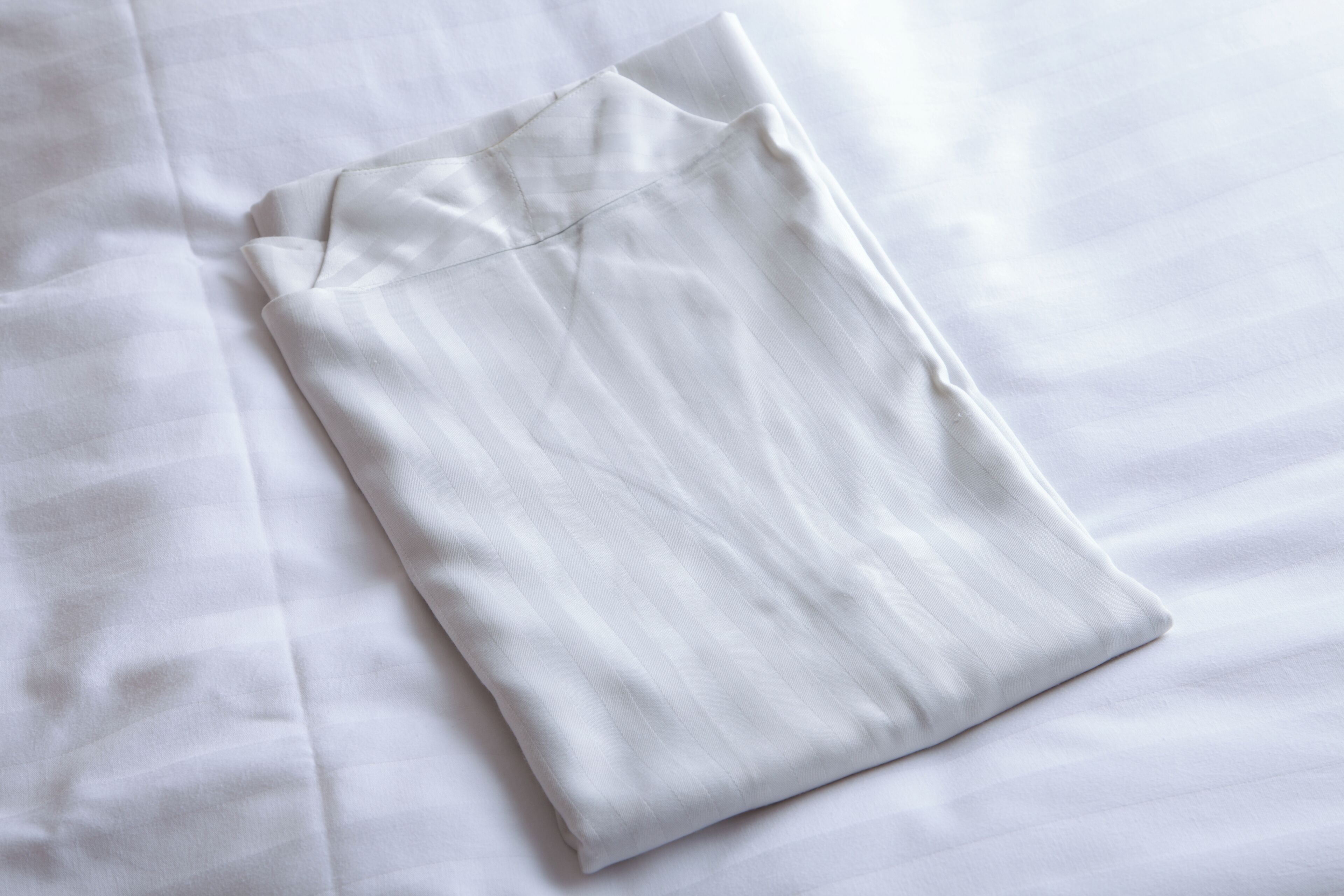 Ein ordentlich gefaltetes, weißes Kleidungsstück auf einem Bett mit weißer Bettdecke.