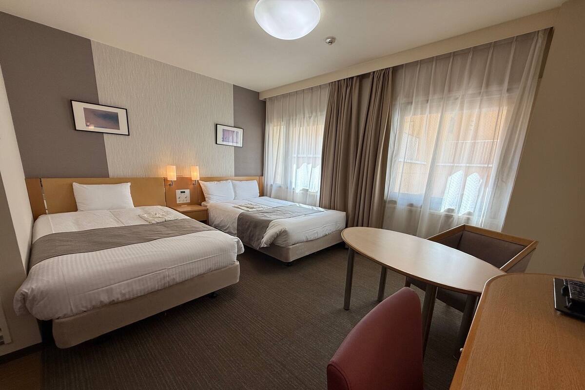 A hotel room with two beds, a desk, and a chair. There are two bedside tables with lamps, a window with curtains, and a wall-mounted picture.