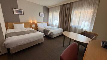 A hotel room with two beds, a desk, and a chair. There are two bedside tables with lamps, a window with curtains, and a wall-mounted picture.
