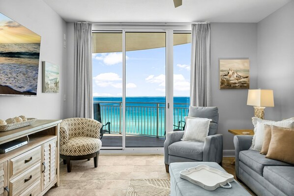 A modern living room with a balcony overlooking a turquoise sea, featuring a sofa, armchair, and a coffee table.