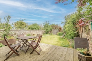A wooden deck with a table and chairs, surrounded by a grassy garden and various plants. - The Whins Cottage | St Andrews (St Andrews)