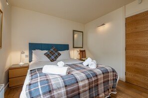 A bedroom with a bed, a nightstand, a bedside table, a wooden wardrobe, and a mirror on the wall. - The Whins Cottage | St Andrews (St Andrews)