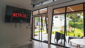 Una habitación moderna con un televisor grande mostrando 'NETFLIX', una puerta corredera de vidrio que da a un patio exterior con sillas y una piscina, y una pequeña mesa con sillas.