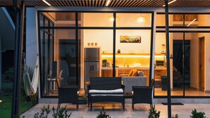A modern house with a glass facade, a dining area with wicker furniture, and a kitchen visible through the glass.