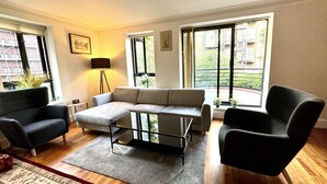Un salon moderne comprenant un canapé, deux fauteuils, une table basse et un lampadaire. De grandes fenêtres offrent une vue sur les bâtiments et la verdure environnante.
