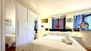 Une chambre avec un grand lit, une armoire, une table de chevet avec une lampe, un miroir et une fenêtre donnant sur des immeubles.
