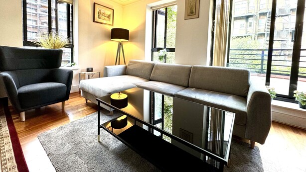 Un salon moderne avec un canapé d’angle, un fauteuil noir, une table basse en verre et un lampadaire. Il y a deux fenêtres avec des rideaux, ainsi qu’une petite table d’appoint avec une lampe.