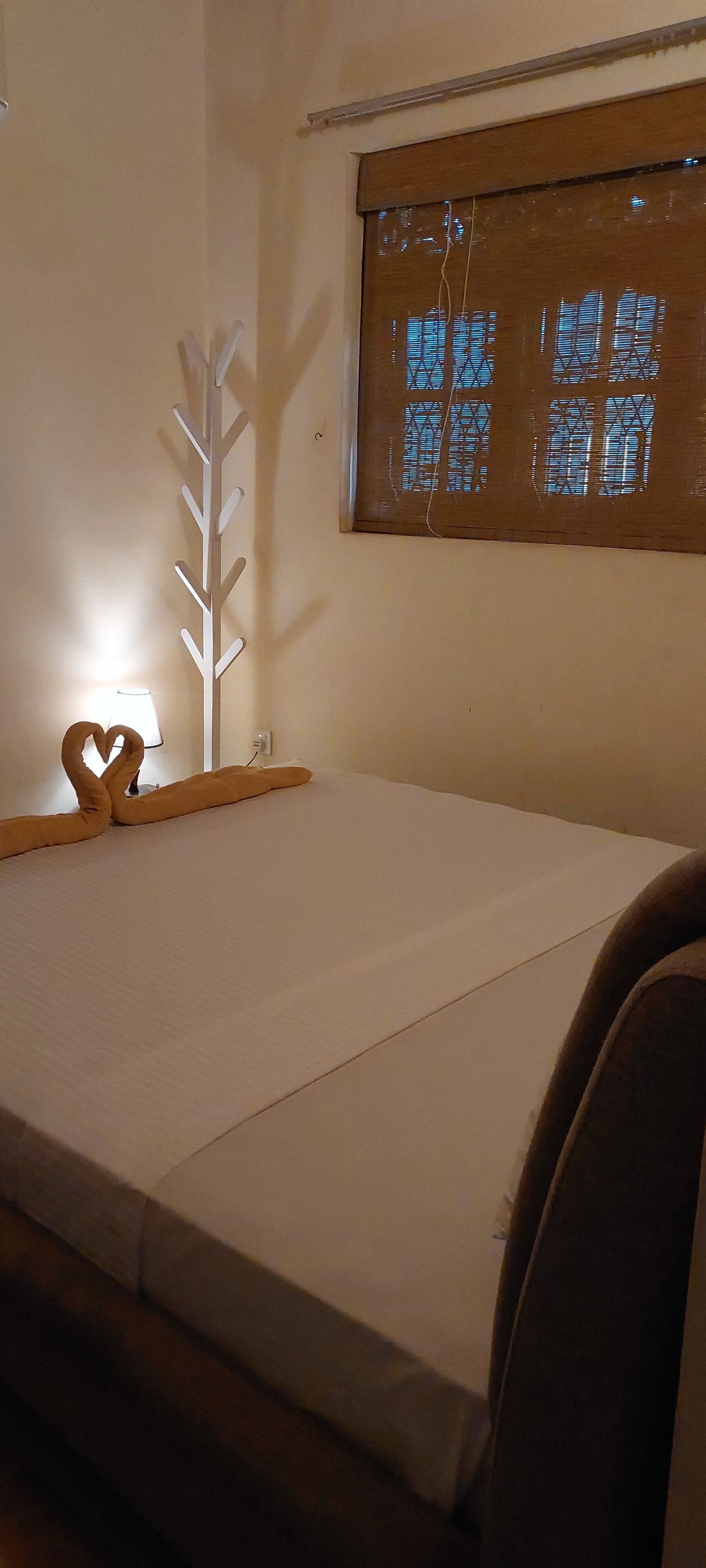 Une chambre avec un lit, une décoration en forme de cœur, une lampe fixée au mur et une fenêtre avec des stores.