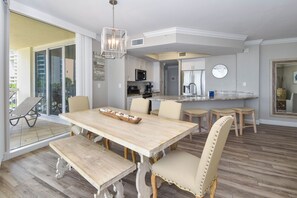A dining area with a wooden table and chairs, a kitchen in the background, and a balcony with a chair.