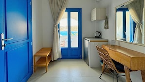 Ein gemütlicher Raum mit blauer Tür, einer kleinen Holzbank, einem Schreibtisch mit Stuhl, einem Minikühlschrank und einem Fenster mit Vorhängen.