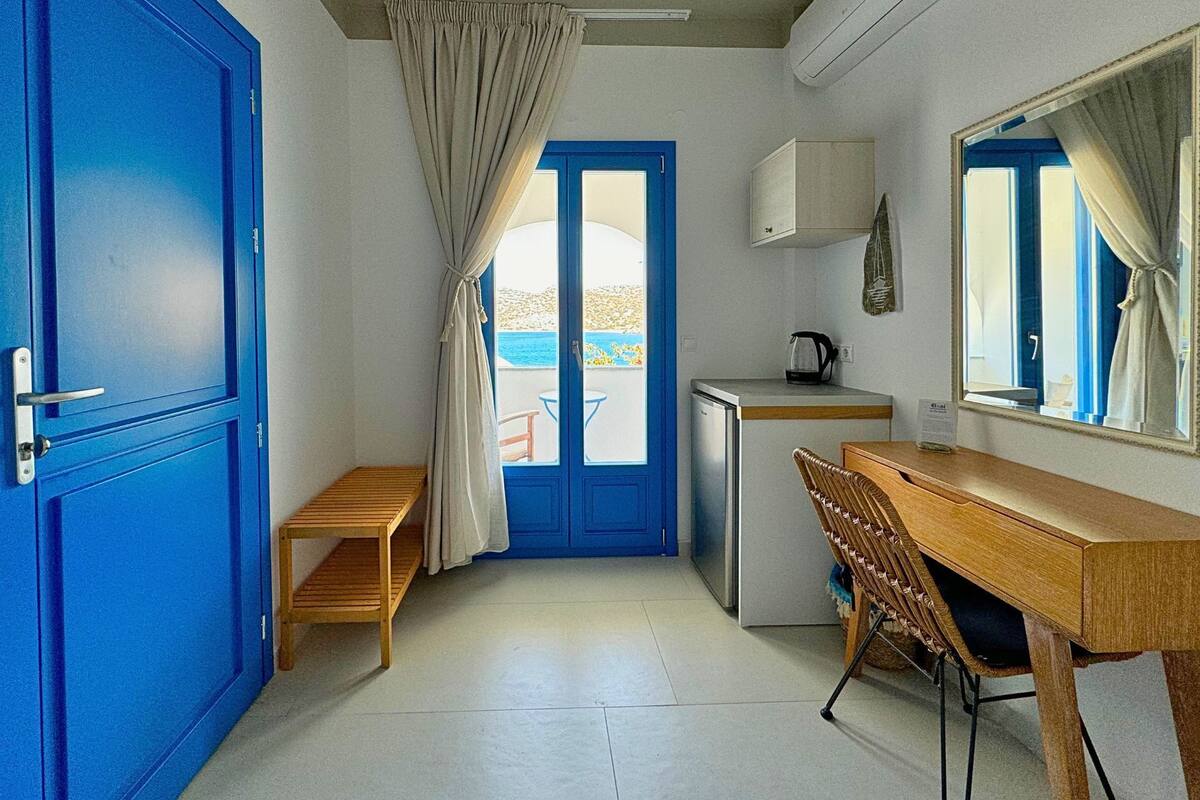 A compact room with a blue door, a small wooden bench, a desk with a chair, a mini-fridge, and a window with curtains.