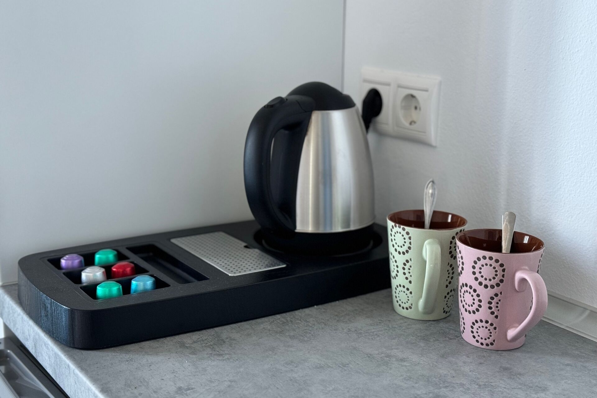 Una encimera de cocina con un hervidor, una bandeja con botellas de colores y dos tazas decoradas.