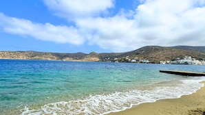 Ein Strand mit klarem türkisblauem Wasser, einem sandigen Ufer und einer Hügellandschaft im Hintergrund mit Gebäuden.