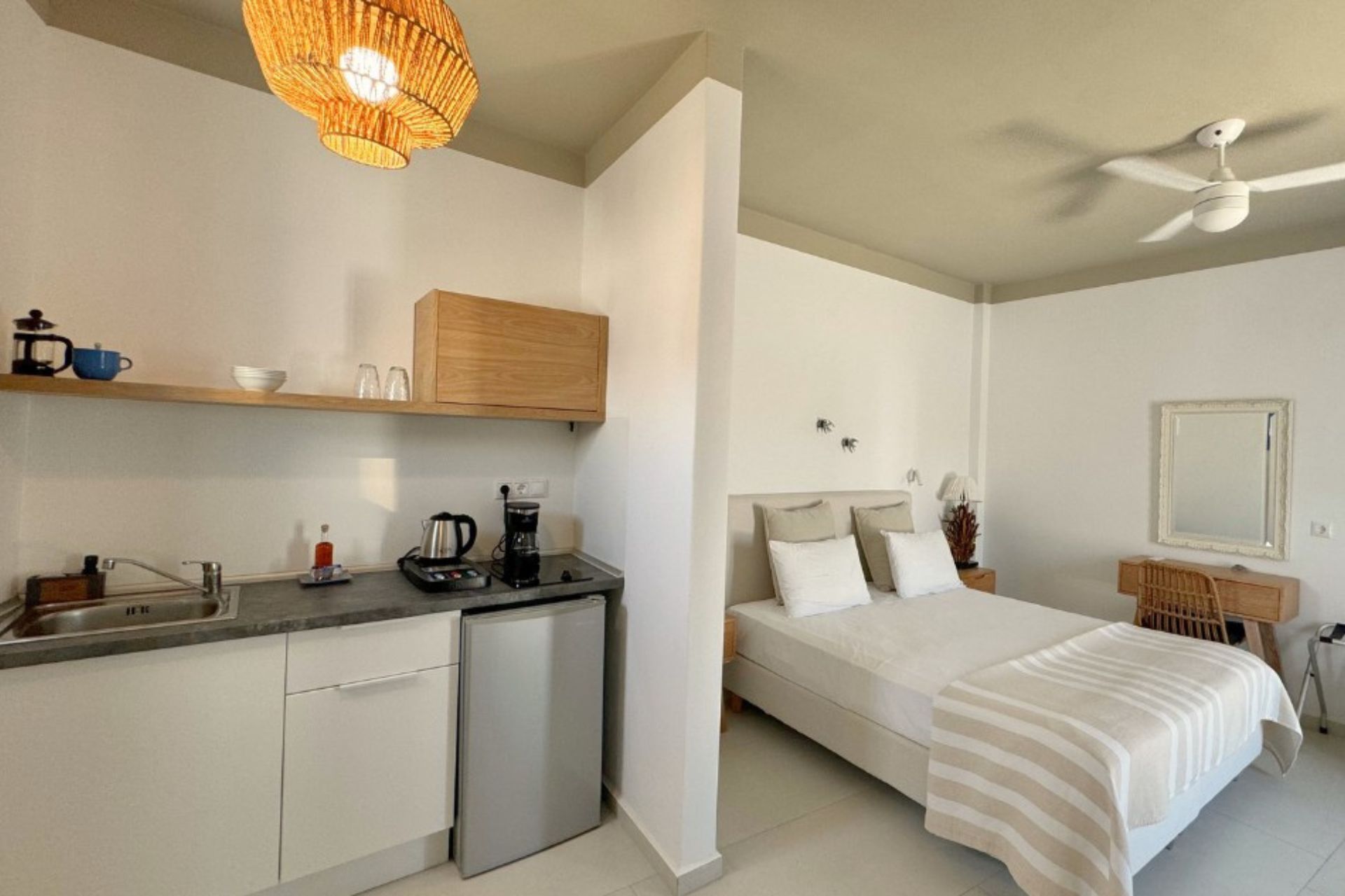 A compact hotel room with a kitchenette, a bed with a striped blanket, a ceiling fan, and a mirror on the wall.