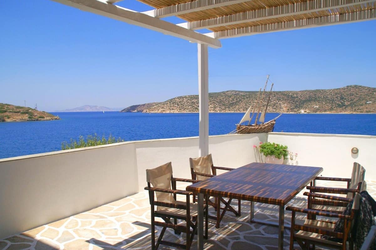 A terrace with a table and chairs, overlooking a blue sea and a hilly landscape.