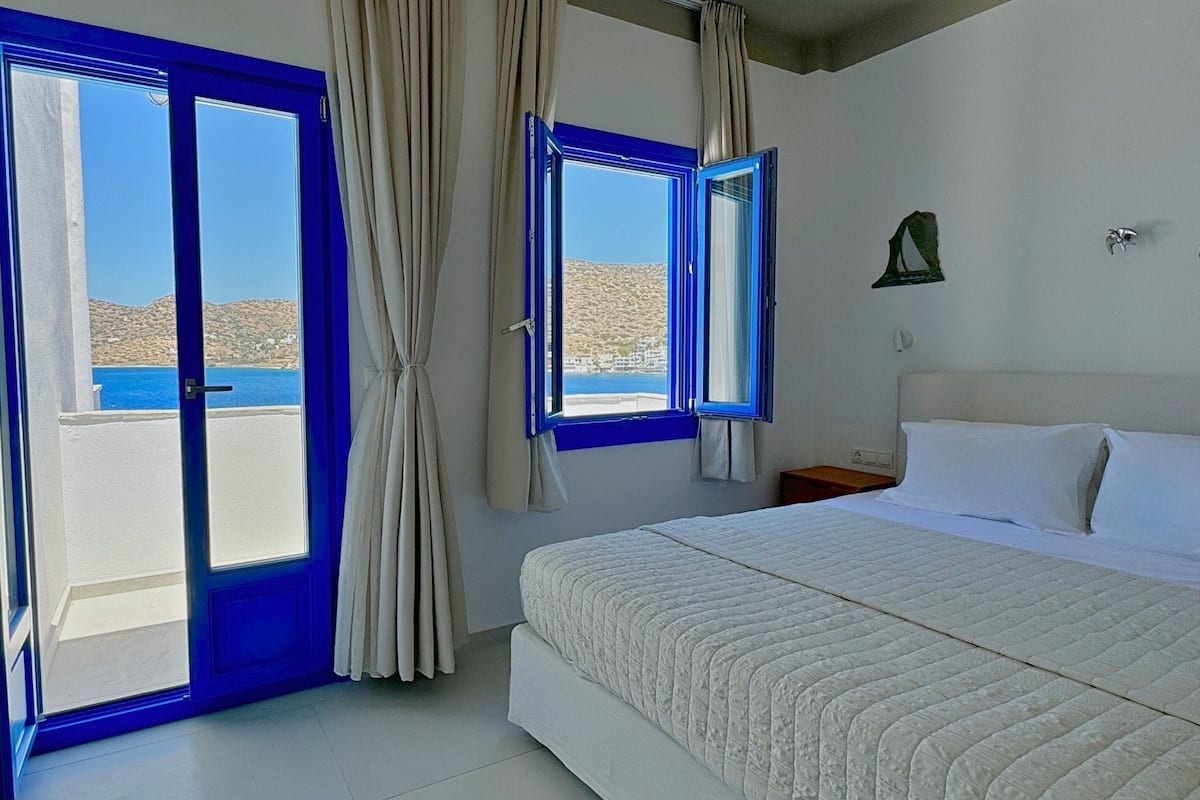 A bedroom with a bed, a nightstand, a window with curtains, and a view of the sea.