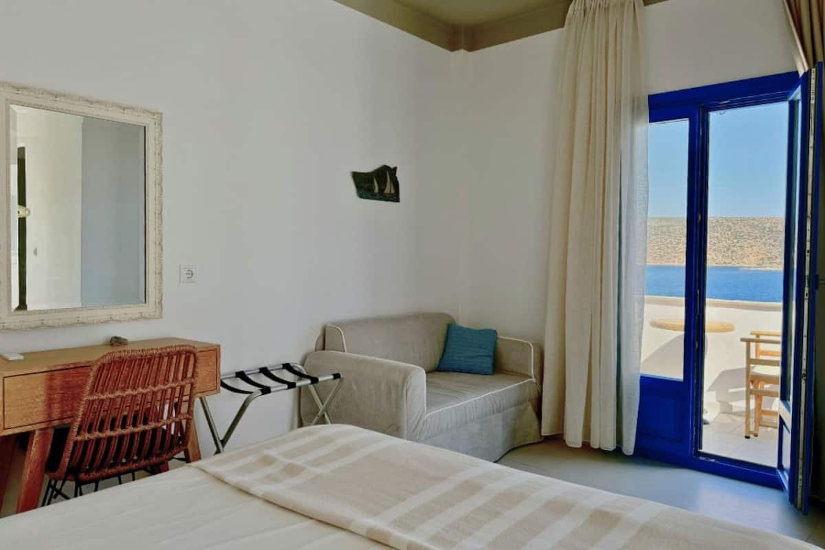 A hotel room with a bed, a desk, a chair, a small table, and a view of the sea through a blue-framed window.