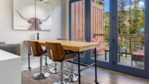 Een moderne eethoek met een houten tafel en metalen stoelen, een groot raam met uitzicht op een terras en bomen, en een aan de muur bevestigd kunstwerk van een stier.