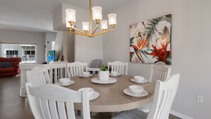 Ein Essbereich mit einem runden Tisch, weißen Stühlen und einer Chandelüster. An der Wand hängt ein Gemälde, im Hintergrund steht ein rotes Sofa.