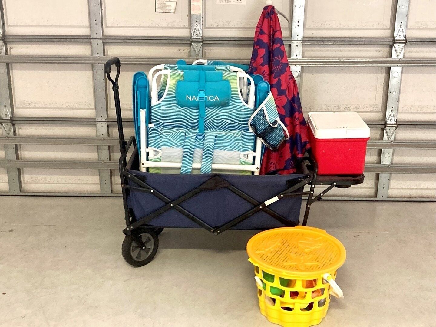 Un carrello bagagli con una borsa a righe blu e bianche con l'etichetta 'NAUTICA', un borsone rosso e un cesto giallo con coperchio forato.