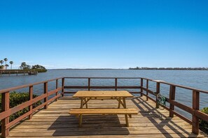 Una terraza de madera con una mesa de picnic y una manguera verde, con vistas a una masa de agua tranquila, tierra distante y palmeras.