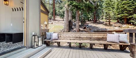 A wooden deck with a bench and cushions, a fireplace, and a view of a forested area.