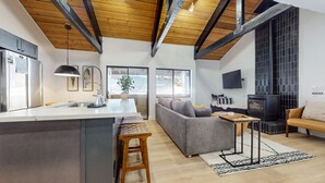A modern kitchen with a central island, a grey sofa, a wooden bench, and a black wood-burning stove. - Bigwood #96 2 Bedroom Condo by RedAwning (Mammoth Lakes)