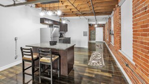 A modern kitchen with a central island, dark wooden flooring, exposed brick walls, and a long hallway with a patterned rug. - Stylish Chic Retreat At Lafayette Lofts 1 Bedroom Apts by RedAwning (Detroit)