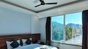 A modern hotel room with a large bed, a ceiling fan, a view of a mountainous landscape through a large window, and a wooden headboard. - HOTEL VAIDIK GANGA  (rishikesh)