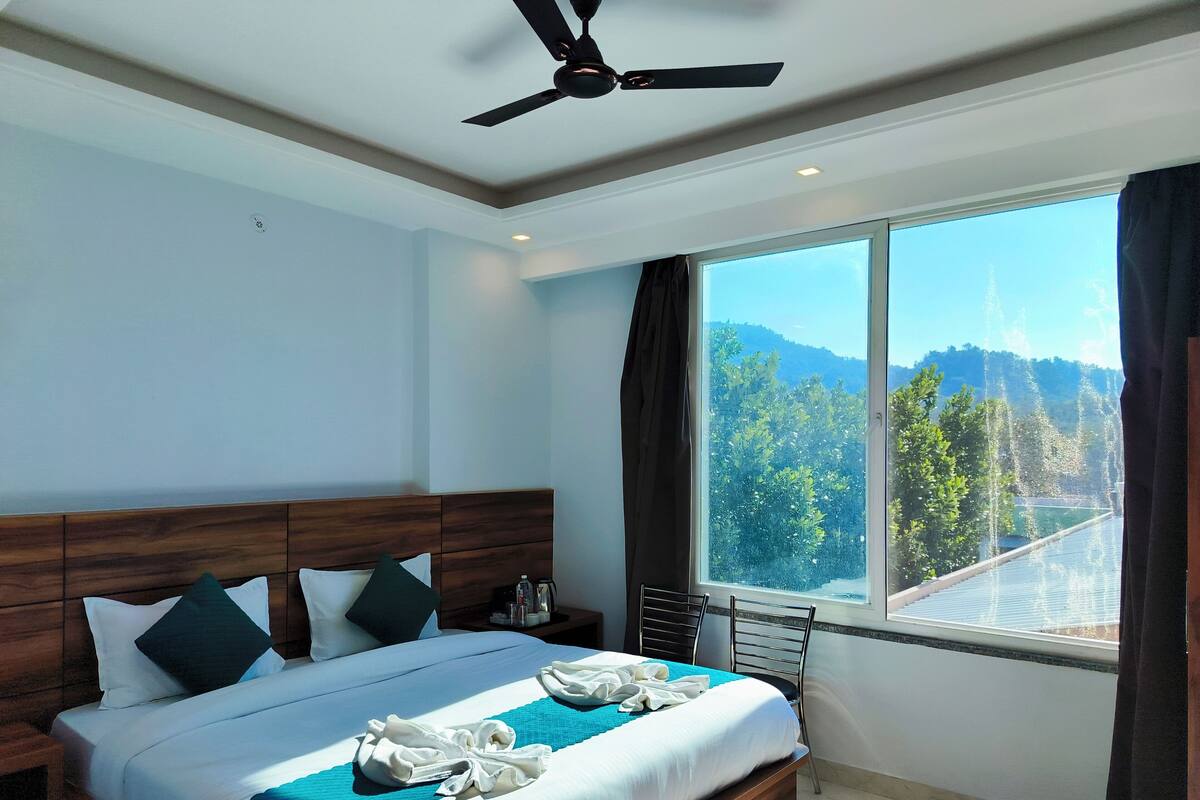 A modern hotel room with a large bed, a ceiling fan, a view of a mountainous landscape through a large window, and a wooden headboard.