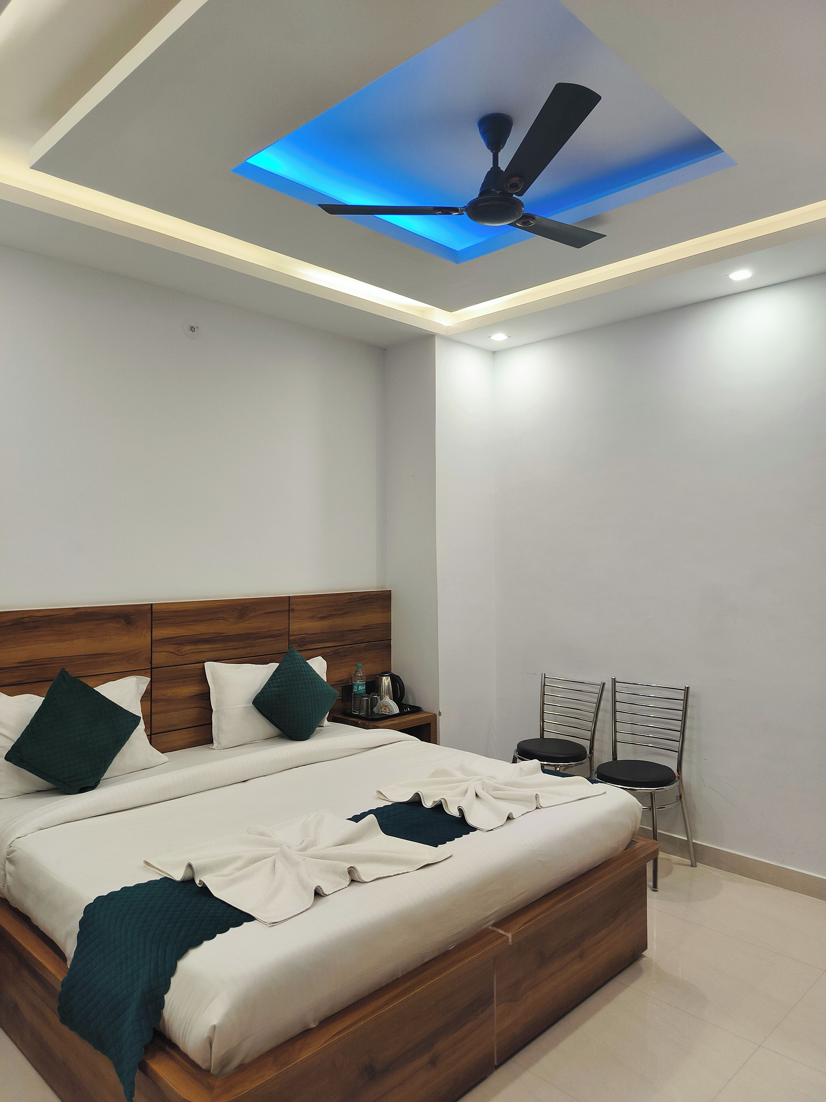 A modern bedroom with a wooden headboard, a bed with white bedding and teal accents, a ceiling fan, and a nightstand with a water bottle and a kettle.