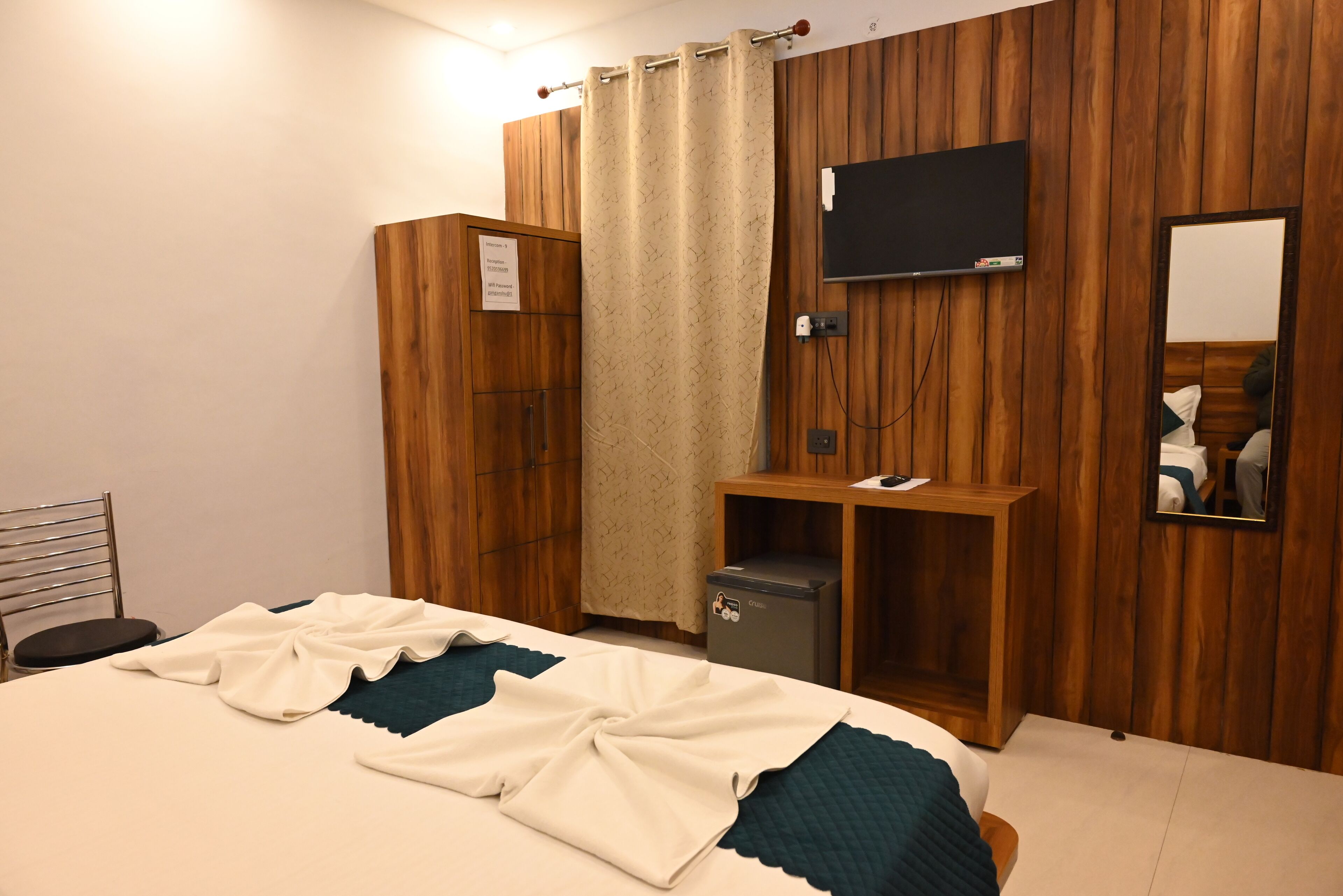 A bedroom with a bed, a TV mounted on the wall, a small desk, a mirror, and a wardrobe.