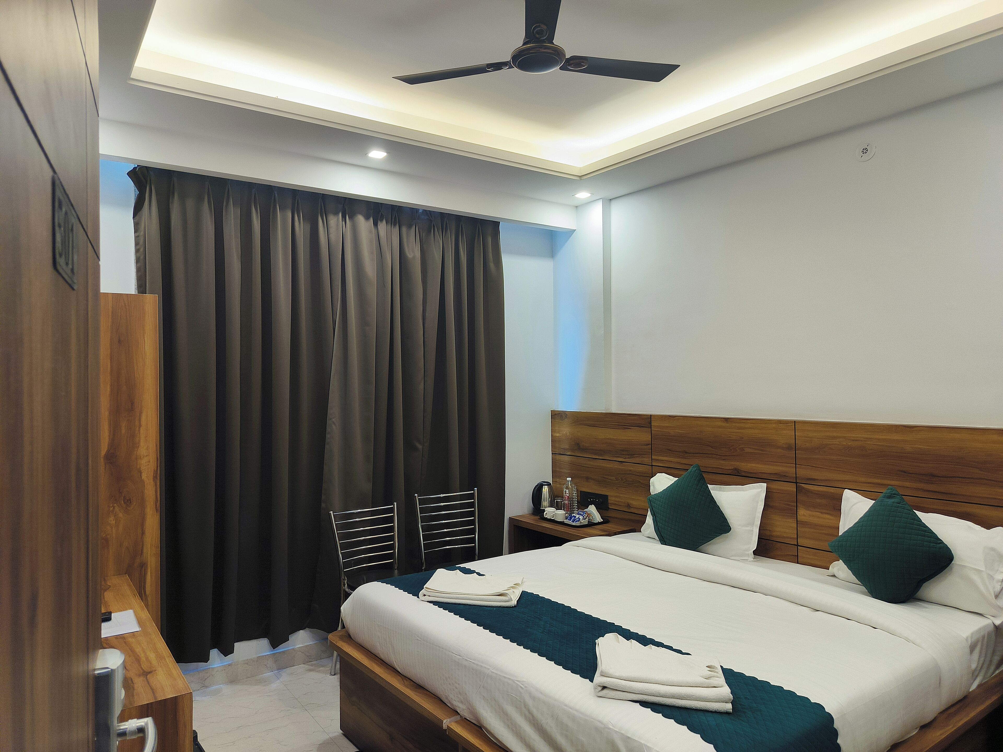 A hotel room with a wooden headboard, a bed with white bedding and green pillows, a nightstand with a phone and a cup, and a ceiling fan.