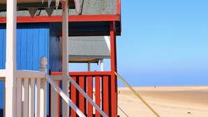 Une maison de plage à l’extérieur rouge et bleu, dotée d’un balcon et offrant une vue sur la plage.