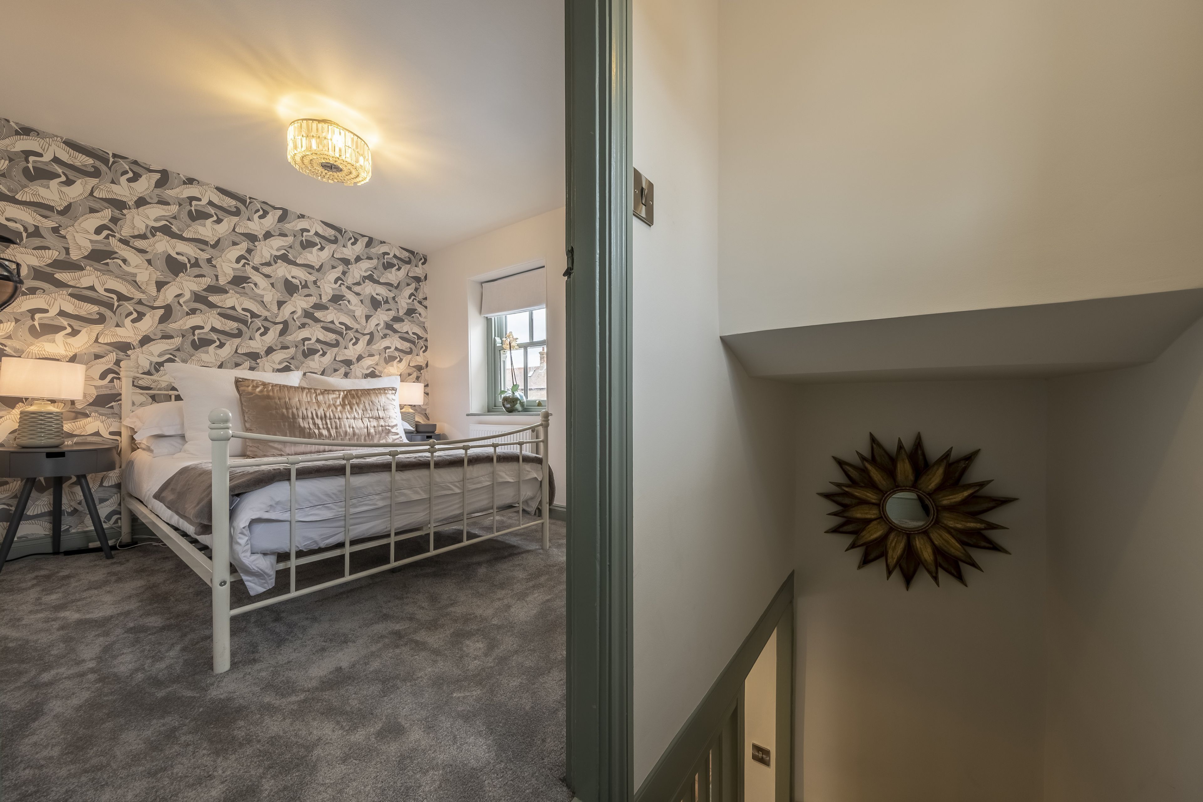 A bedroom with a bed, a nightstand, a lamp, a wall with a patterned wallpaper, a window with a view, and a decorative mirror on the wall.