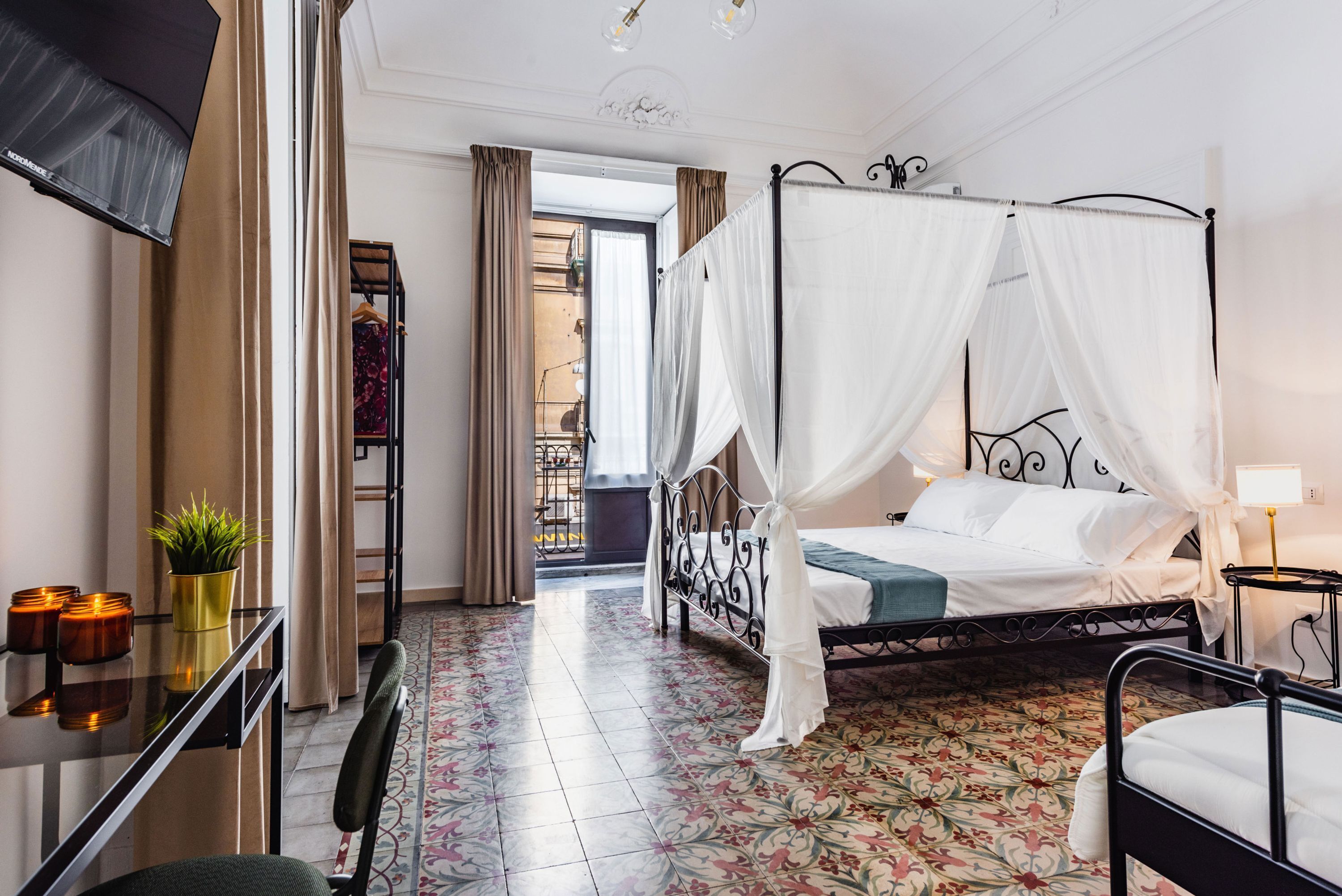A bedroom with a four-poster bed, a desk with a chair, a potted plant, and a nightstand with a lamp.