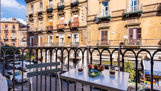 Un balcon avec une table, des chaises et une vue sur une rue bordée de voitures garées et d’immeubles.
