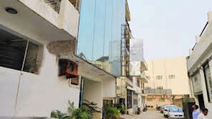 A narrow street with buildings on both sides, some with balconies. There are cars parked along the street, and a few people are visible. - HOTEL SMART STAY (New Delhi)