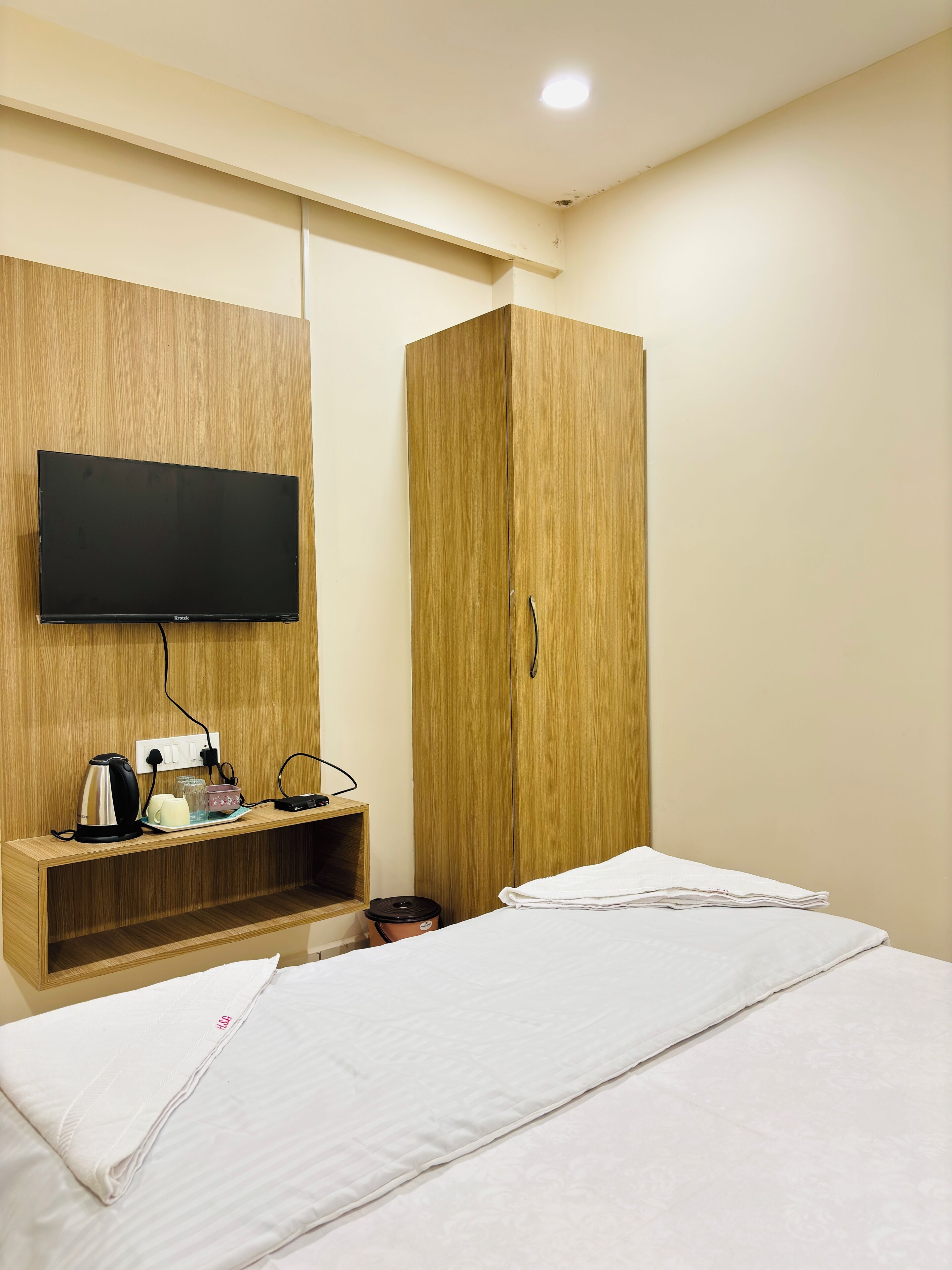 A hotel room with a bed, a wooden wardrobe, a TV mounted on the wall, and a small shelf with a kettle and glasses.