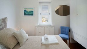 A bedroom with a bed, a chair, a wardrobe, a window with a view, and an air conditioning unit.