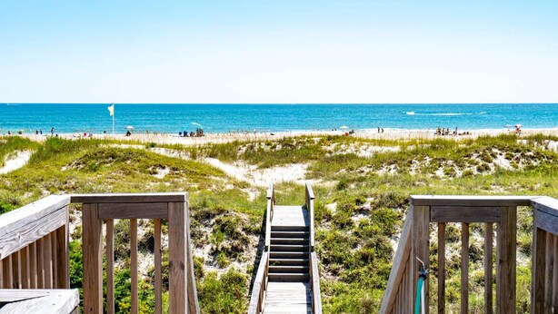 Eine Holztreppe führt zu einem Strand mit freiem Blick auf das Meer und Personen in der Ferne.