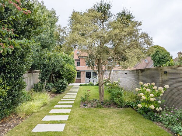 Stroll through the serene garden, surrounded by lush plants and shrubs, to the charming residential property with a red-tiled roof.