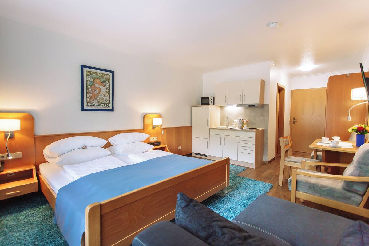 Quarto de hotel com cama, mesas de cabeceira, escrivaninha, cadeira, cozinha compacta e banheiro.